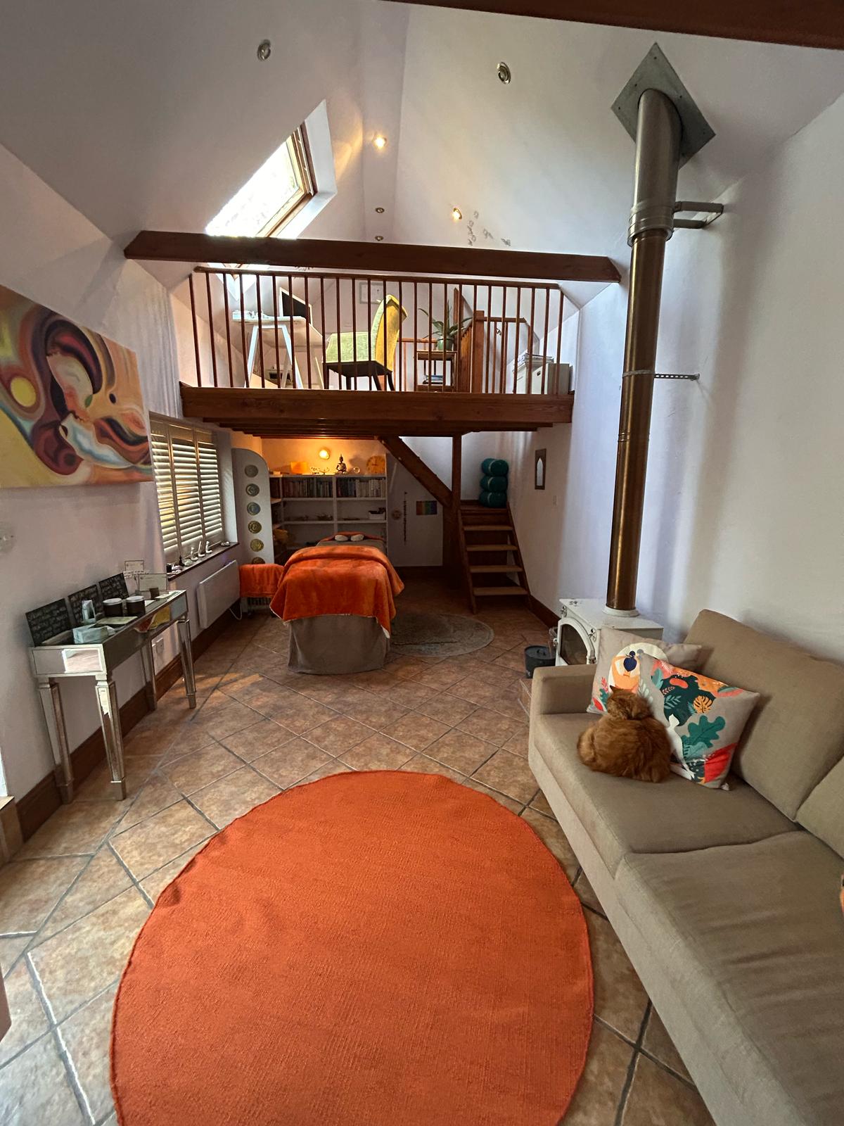 The treatment couch dressed in an orange blanket, a lit lamp, a bookshelf of oils and small objects, shutters, and the wooden stair behind.