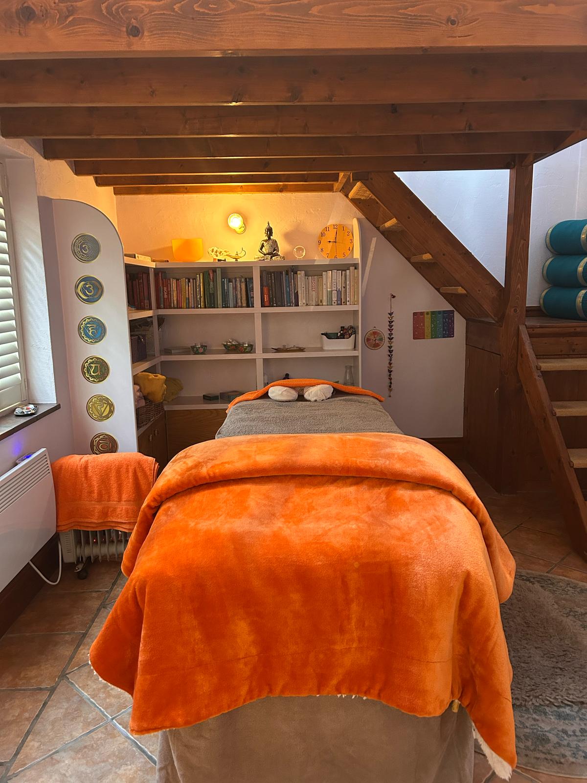Faren’s treatment room at The Coach House: a beamed, white-plastered room with a mezzanine gallery, a wood-burner flue, a round terracotta rug on a tiled floor and the treatment couch under an orange blanket.